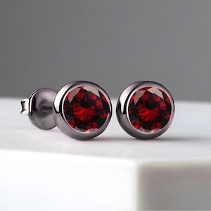 May include: A pair of stud earrings featuring round, deep red gemstones set in dark gray metal. The earrings have a simple, elegant design, with the gemstones catching the light. The background is a soft gray, highlighting the jewelry.