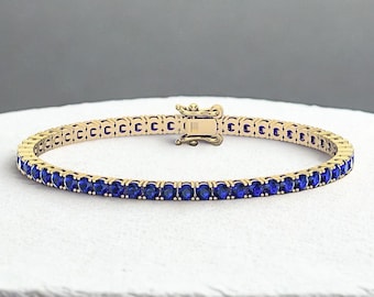 May include: A gold-toned tennis bracelet featuring a series of oval-cut, deep blue gemstones. The bracelet has a secure clasp and is displayed on a white surface. The gemstones are evenly spaced, creating a continuous line of color.