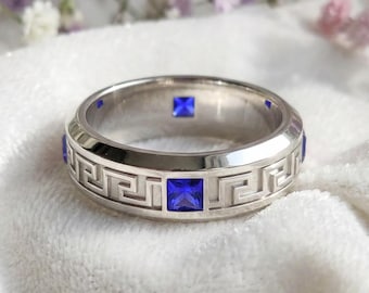 May include: A silver ring with a Greek key design and square blue gemstones. The ring has a polished finish and is set with multiple blue stones around the band. The ring is displayed on a white, textured surface.