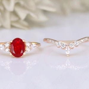 May include: A set of two gold rings featuring a large red oval gemstone and smaller clear gemstones. The engagement ring has a single large red oval gemstone surrounded by smaller clear gemstones. The wedding band is a delicate curved band with clear gemstones.