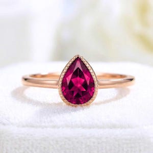 Pode incluir: Um anel de ouro rosa com uma pedra preciosa rosa-escura em forma de pera, engastada num bisel com detalhes milgrain. A banda é lisa e polida, apresentando um design simples que destaca a pedra central facetada e a sua montagem metálica.