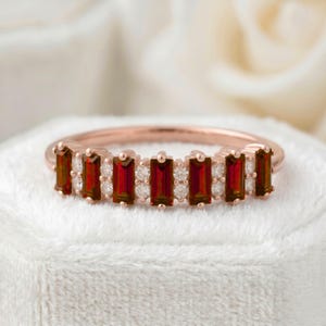 Puede incluir: Anillo de oro rosa con piedras rojas rectangulares alternadas con pequeñas piedras claras redondas. El anillo tiene una banda delicada y un patrón repetitivo de piedras en la parte superior, creando un diseño brillante y elegante.