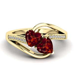 May include: A gold-colored ring featuring two heart-shaped, deep red gemstones. The band is adorned with small, clear gemstones. The ring's design is elegant and romantic, perfect for a special occasion.
