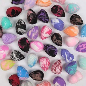May include: A collection of teardrop-shaped makeup sponges in various colors, including purple, pink, orange, and blue. Each sponge features a marbled design, creating a unique pattern. The sponges are arranged on a white surface.