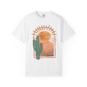 May include: A white short-sleeved t-shirt featuring a graphic print of a green cactus standing before a large orange sun. The design is framed by a pink arch with radiating lines and includes the text Lottie Louise in a cursive script font.