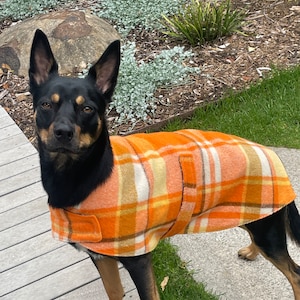 May include: A black and tan dog wearing an orange, yellow, and white plaid dog coat. The dog is standing on a wooden deck, with a garden and rocks in the background. The coat has a classic, tailored design.