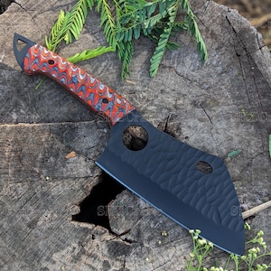 May include: A black cleaver with a red and grey patterned handle. The cleaver has a textured blade and two holes in the blade. The cleaver is sitting on a tree stump.