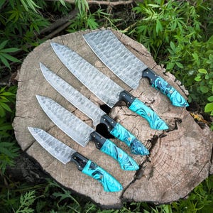 May include: A collection of six handcrafted kitchen knives with textured blades and turquoise and black handles. The knives are arranged on a weathered wooden surface, showcasing their unique design and craftsmanship. The blades have a distinctive pattern.