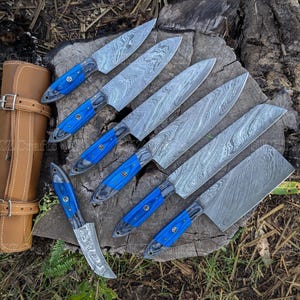 May include: A set of seven Damascus steel knives with blue handles, arranged on a wooden surface. The blades have a distinctive pattern. A brown leather knife roll is visible on the left side.
