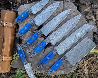 May include: A set of seven Damascus steel knives with blue handles, arranged on a wooden surface. The blades have a distinctive pattern. A brown leather knife roll is visible on the left side.
