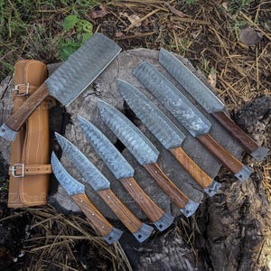 May include: A collection of handcrafted knives with wooden handles and patterned blades, arranged on a tree stump. A leather sheath is visible on the left. The knives vary in size and shape, with a cleaver and several smaller blades.