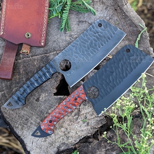 May include: Two cleaver knives with textured blades and handles. One knife has a black blade and a red and white patterned handle. The other has a grey blade and a blue handle. A leather sheath is visible in the background.