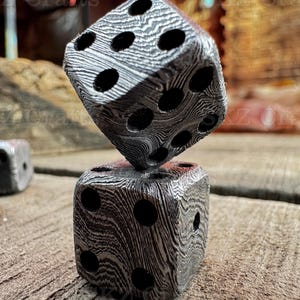 May include: Two six-sided dice stacked vertically, featuring a distinct black and silver layered pattern resembling Damascus steel. Each die has circular indentations representing the pips, with a third die partially visible in the background.