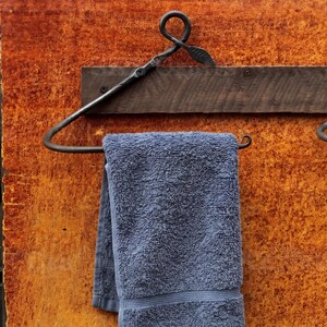 Pode incluir: Um toalheiro de metal cinza-escuro com estrutura triangular e um detalhe decorativo de folha no topo, montado numa tábua de madeira horizontal. Uma toalha de felpa azul dobrada está pendurada na barra inferior do suporte metálico.