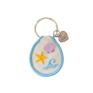 Puede incluir: Un llavero con temática de playa con un diseño bordado blanco en forma de lágrima. Presenta una estrella de mar amarilla, una concha morada y una ola azul. El diseño está delineado con pespuntes azul claro. Un dije de corazón plateado y un llavero completan el accesorio.