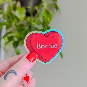 Könnte beinhalten: Ein roter herzförmiger Aufkleber mit holografischem Finish. Der Aufkleber sagt "Bite me" in weißer Schrift.