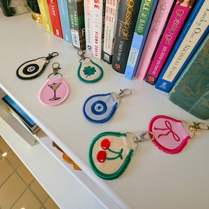 Op de afbeelding: Een verzameling geborduurde sleutelhangers met diverse ontwerpen, waaronder een cocktailglas, een klavertjevier, kersen, een 8-ball, een oog en de letter "R". Elke sleutelhanger heeft een metalen sluiting.