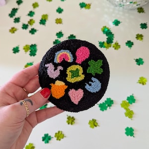 Könnte beinhalten: Ein schwarzer runder Nadelfilz mit bunten gestickten Designs, darunter ein Regenbogen, ein Stern, ein Herz, ein Vogel, ein Topf und eine Blume.