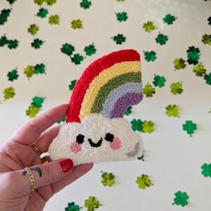 Puede incluir: Una nube blanca con una cara sonriente y mejillas rosadas, coronada por un arcoíris hecho de hilo rojo, naranja, amarillo, verde, azul y morado.