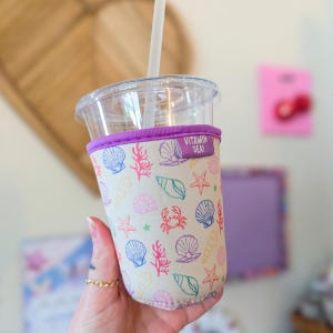 Puede incluir: Un vaso de plástico transparente con pajita y una funda de neopreno beige con un patrón colorido de conchas, estrellas de mar, corales y cangrejos. La funda tiene un borde morado y una etiqueta de tela que dice VITAMIN SEA! en blanco.