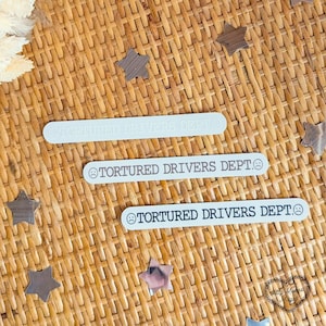 May include: Three white rectangular stickers with rounded edges, each bearing the text "TORTURED DRIVERS DEPT" in a sans-serif font. The middle sticker includes a sad face emoji. The stickers are on a woven surface with star-shaped confetti.