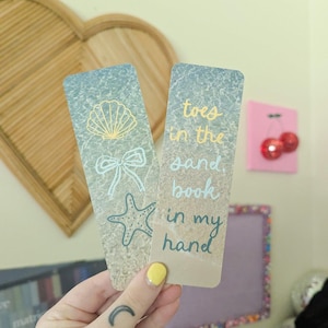 May include: Two beach-themed bookmarks. One features a seashell, bow, and starfish. The other reads "toes in the sand, book in my hand." The bookmarks have a sand and sea design with blue and yellow accents.