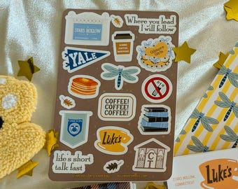 May include: A sticker sheet featuring various designs and text, including "Stars Hollow," "Yale," and "Luke's." The stickers are brown with blue, white, and yellow accents. Other stickers include a coffee cup, pancakes, and a dragonfly.