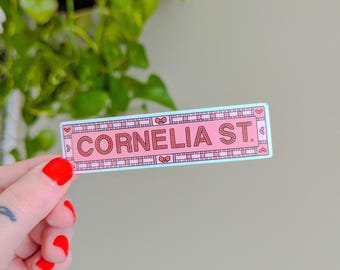 May include: A pink and red sticker with the text "CORNELIA ST." in a white box with a checkered pattern. The sticker has a heart-shaped border.