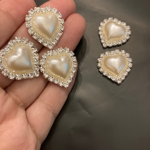 May include: Several heart-shaped embellishments with a pearl-like center and a sparkling rhinestone border. The hearts vary in size and are displayed against a dark background. These decorative elements could be used for crafts or fashion.