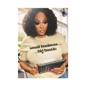 May include: A woman wearing a beige t-shirt with the text "small business... ...big hustle" in black. She is holding a black tablet computer and looking at the screen.