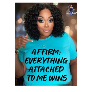 May include: A woman wearing a turquoise blue t-shirt with the text "AFFIRM: EVERYTHING ATTACHED TO ME WINS" printed in black.