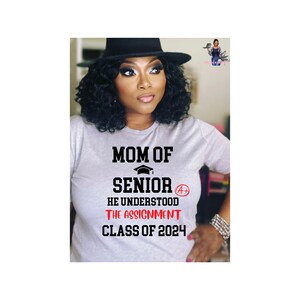 May include: A woman wearing a gray t-shirt with the text "Mom of Senior He Understood The Assignment Class of 2024" and a black hat.