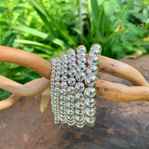May include: A stack of silver bead bracelets, each with a different size bead. The bracelets are arranged in a circle on a wooden branch.