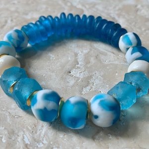 May include: A blue and white glass bead bracelet with gold accents. The bracelet features a variety of different shaped beads, including round, square, and rectangular beads.