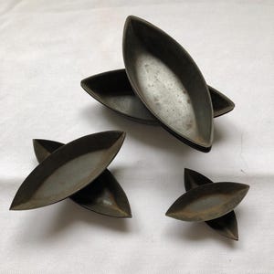 May include: Several dark metal leaf-shaped dishes are arranged on a white surface. The dishes have a slightly textured appearance and a dark, metallic color. The arrangement includes stacks and individual pieces.