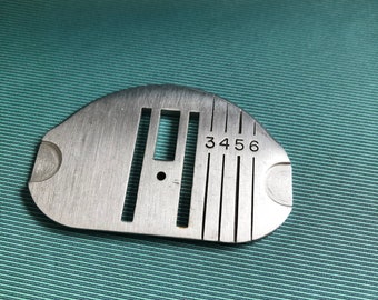 May include: A silver sewing machine foot with a series of grooves and a small hole. The numbers 3, 4, 5, and 6 are engraved on the foot.