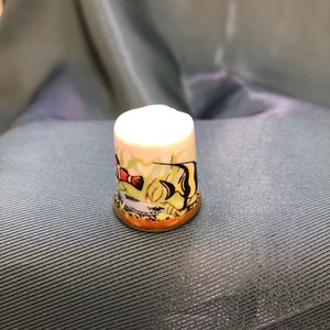 May include: A white porcelain thimble with a gold rim. The thimble is decorated with a colorful painting of two fish swimming in a coral reef.