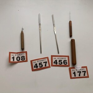 Könnte beinhalten: Eine Sammlung von Häkelnadeln und Werkzeugen, darunter Häkelnadeln aus Holz und Metall sowie ein Holzetui. Die Werkzeuge sind auf einer weißen Oberfläche angeordnet, mit orangefarbenen und weißen Preisschildern.