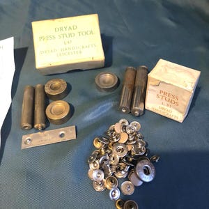 May include: A set of vintage metal press studs and a tool for attaching them. The tool is in its original box, labeled "Dryad Press Stud Tool L47 Dryad Handicrafts Leicester". The box of studs is labeled "Press Studs L 83 Dryad Handicrafts".