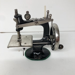 May include: A vintage black and silver Singer hand-crank sewing machine featuring a metal base, a needle bar, a presser foot, and a side-mounted hand wheel with a wooden handle. The brand name SINGER is clearly printed in gold on the black frame.