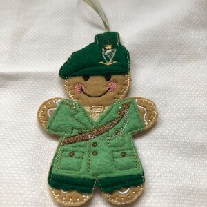 May include: A felt gingerbread man ornament wearing a green uniform and beret. The ornament has a smiling face with pink cheeks and a light brown body. The uniform has gold accents and a brown belt. A gold ribbon is attached for hanging.