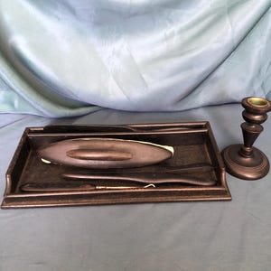 May include: A black wooden tray with a black leather lining holds a set of antique shaving tools, including a straight razor, a strop, and a shaving brush. A black wooden candle holder with a green base sits to the right of the tray.