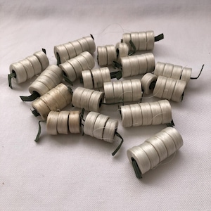 May include: A group of off-white cylindrical objects, possibly spools or decorative elements, are arranged on white fabric. Each cylinder is wrapped with a fine, light-coloured material and has a small green piece attached. The objects vary slightly in colour.