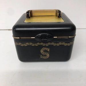 May include: A black rectangular storage case with a gold-coloured top handle and latch. The front features a gold decorative scroll pattern border and a central emblem with the text SINGER SEWING MACHINES printed in black on a gold background.