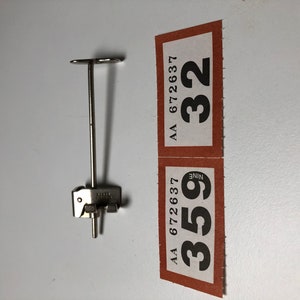 May include: A silver metal clamp with a long, thin rod extending from the top. The clamp has a small, round knob on the side. The rod has a T-shaped top. The clamp is labeled with the numbers "359" and "32" and the letters "AA 672637".