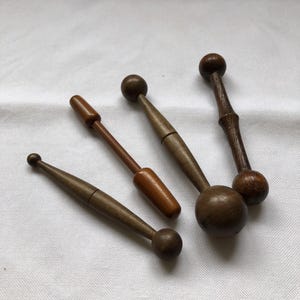 May include: Four wooden tools with rounded ends and tapered handles. The tools vary in color from light to dark brown. The objects are arranged on a white, textured surface.