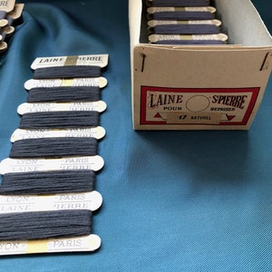 May include: A collection of navy blue thread spools labeled "LAINE PIERRE" and "LYON PARIS" are displayed. The spools are arranged in a cardboard box and in a row. The box has a red and white label with the text "LAINE POUR REPRISER".