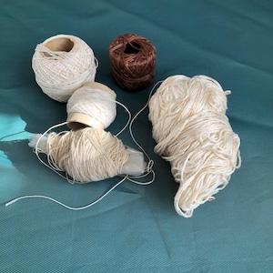 May include: Four spools of thread, two white, one brown and one beige, on a teal surface.