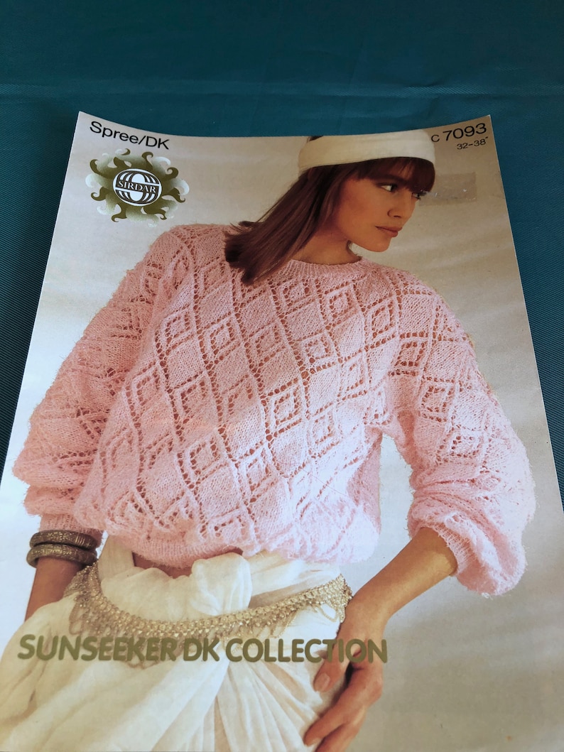 Pode incluir: Um su&eacute;ter de malha rosa com um padr&atilde;o de diamantes. O su&eacute;ter tem mangas compridas e uma gola redonda. A modelo est&aacute; usando uma faixa de cabe&ccedil;a branca e uma saia branca. A imagem inclui o texto "Spree/DK", "C 7093 32-38", e "SUNSEEKER DK COLLECTION".