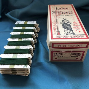 May include: A stack of green embroidery floss spools labelled "Laine St Pierre" in a white box with gold lettering. The spools are arranged in a row and are ready to be used for sewing or embroidery projects. The box is labelled "Laine St Pierre Pour Repriser" and "M-M-Lyon".
